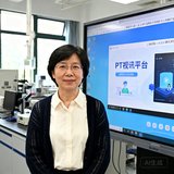 上海某双一流大学计算机学院陈雅芳教授的半身照，她穿着米白色衬衫外搭深色针织开衫，站在实验室大屏幕前，屏幕上正运行着PT视讯平台的远程教学界面。